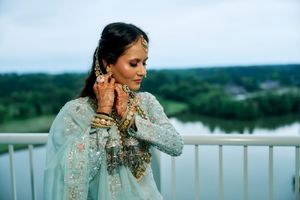South Asian bride in light blue lehenga adjusting earrings on balcony with henna hands and gold jewelry.
