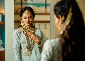 Indian bride in light blue lehenga admires her gold jewelry and maang tikka in mirror during bridal preparation.