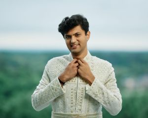 Young man in white embroidered sherwani adjusting collar outdoors with lush green landscape behind him.
