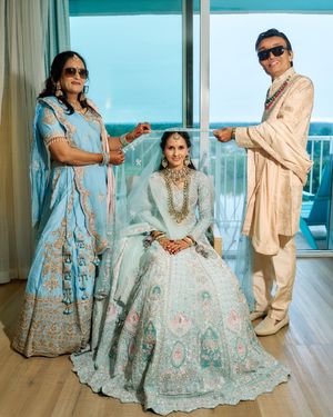 Indian bride in light blue lehenga seated while parents hold her dupatta near a window with ocean view.