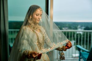 South Asian bride in embroidered lehenga and veil holding kalire on a balcony.