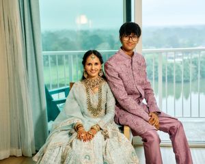 Indian bride in light blue lehenga with gold jewelry sits beside young man in pink sherwani by scenic window.