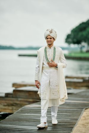 Indian groom in white sherwani and turban standing on wooden dock by water.