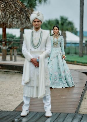 Indian groom in white sherwani and turban stands before bride in light blue embroidered lehenga at outdoor wedding.