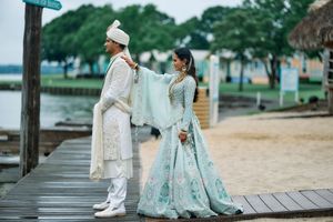 Indian couple in traditional wedding attire on a wooden dock, bride in mint lehenga adjusting groom's white sherwani.