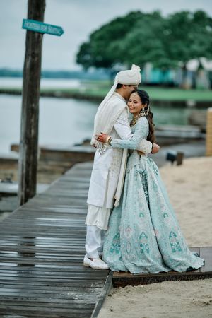 Indian couple embracing on a wooden dock, groom in white sherwani and bride in mint green lehenga by the water.
