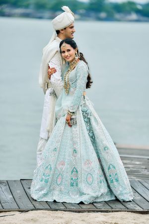 Indian bride and groom posing on a waterfront dock, bride in embroidered mint lehenga with gold jewelry.