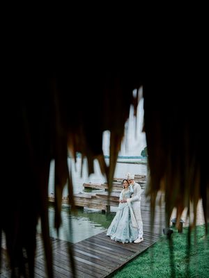 Indian wedding couple embraces on wooden dock framed by dark foliage with serene lake backdrop.
