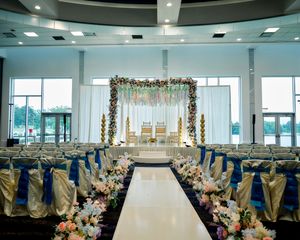 Elegant Indian wedding ceremony hall with floral mandap, blue sash chairs, and white aisle runner.