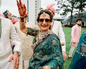 Indian woman in teal saree with mehndi hands raised, wearing sunglasses and traditional gold jewelry.