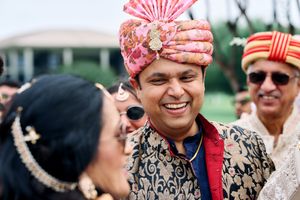 Smiling Indian groom in ornate gold sherwani and pink floral turban during wedding baraat ceremony.