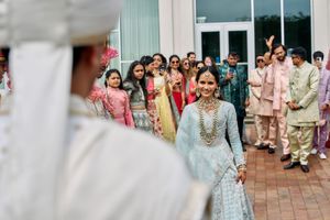 Bride in light blue lehenga with gold jewelry walks toward groom during Indian wedding ceremony outdoors.