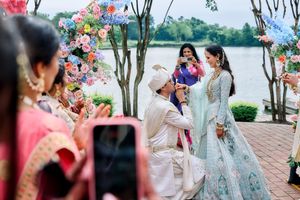 Groom kneeling before bride at outdoor Indian wedding ceremony under a floral arch by the lake.