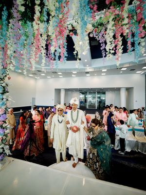 Indian wedding ceremony with groom in white sherwani walking under floral ceiling decor at elegant venue.