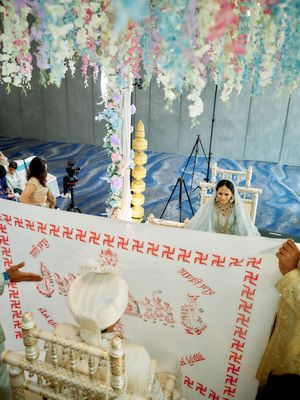 Traditional Indian wedding ceremony with ornate white fabric held up, floral decorations, and bride in blue bridal attire.