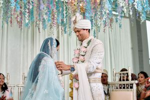 Indian groom in white sherwani placing ring on bride in light blue lehenga under floral canopy.