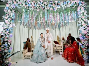 Indian wedding couple under floral arch with pink and blue flowers at ceremony.