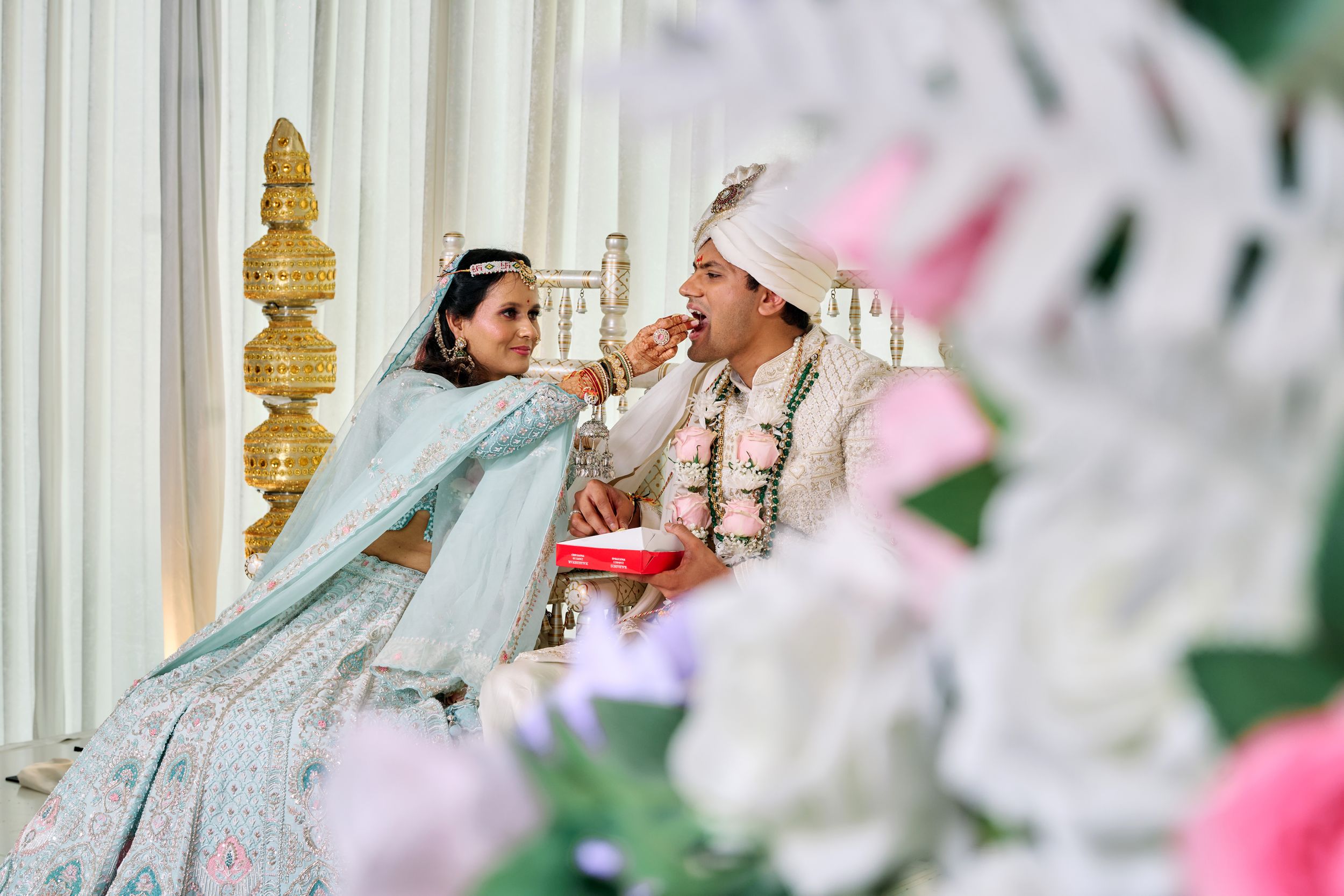 Bride in blue lehenga feeds groom during traditional Indian wedding ceremony with floral decorations.