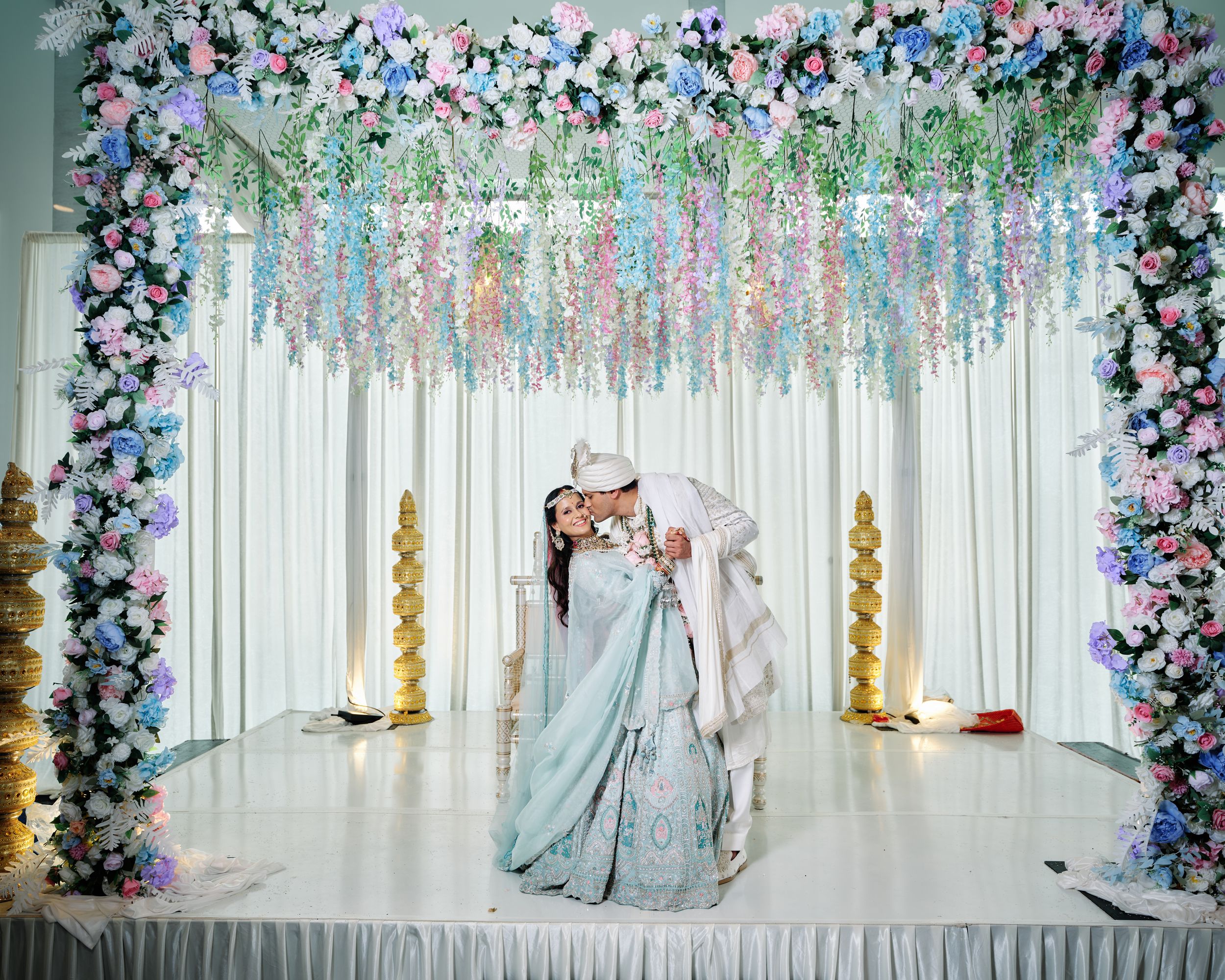 Bride and groom share a romantic moment under a floral arch with pink and white flowers at an Indian wedding.