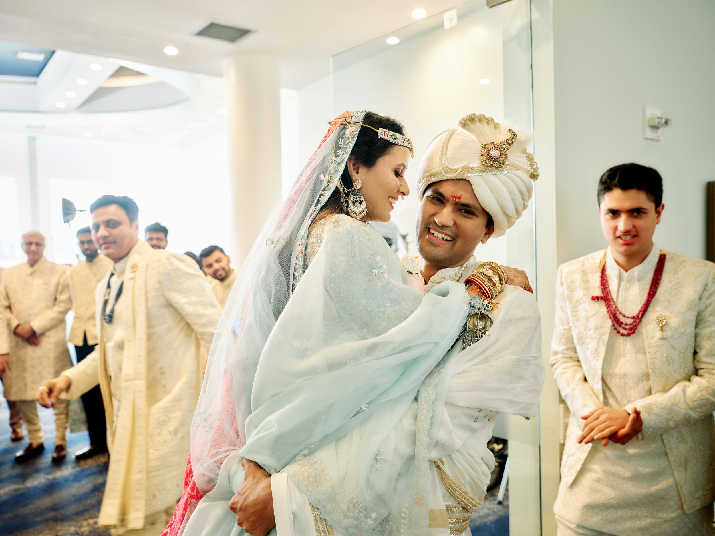 Joyful Indian wedding moment with groom in white sherwani lifting bride in traditional attire surrounded by guests.
