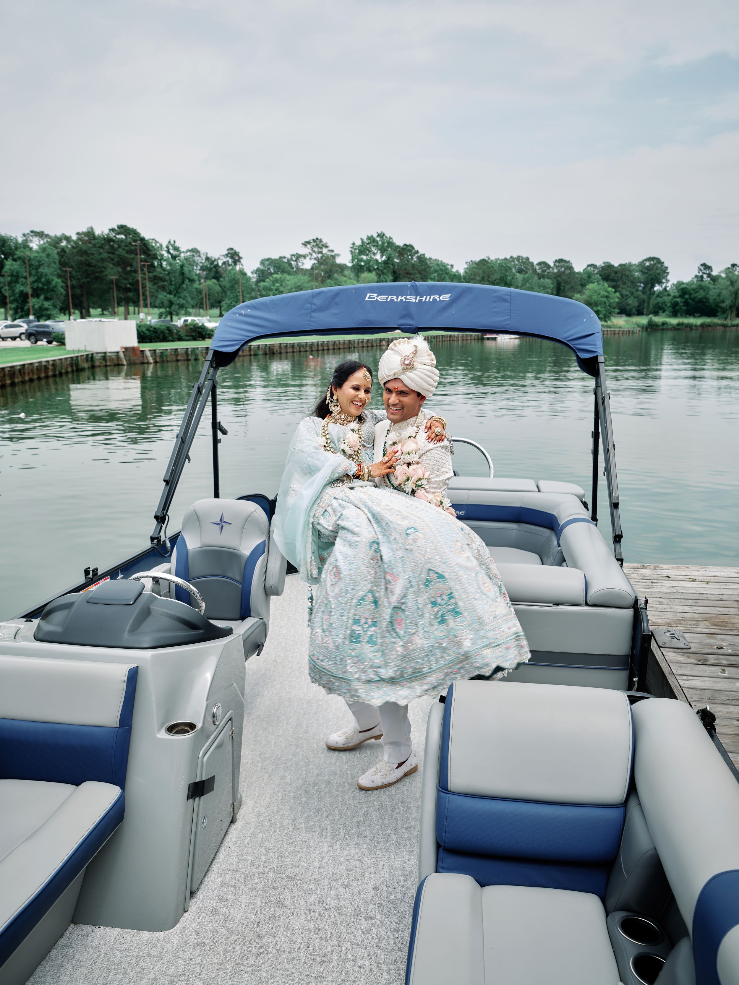 Indian wedding couple in traditional attire posing on a blue pontoon boat docked on a serene lake.