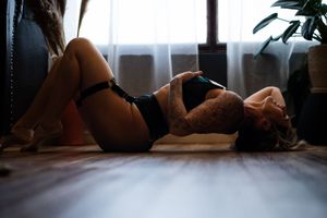 Woman in black lingerie lying on hardwood floor in moody boudoir setting with soft window light and plants.