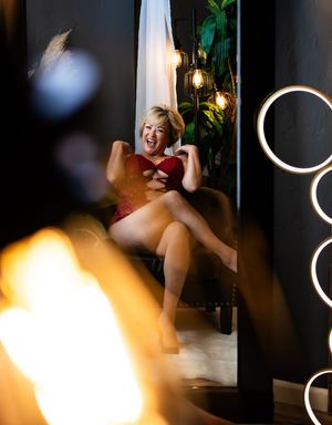 A laughing blonde woman in red lingerie poses playfully in a glamorous studio with circular lights and lush greenery.