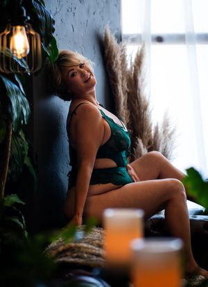 Smiling blonde woman in green lace lingerie posing against a dark wall surrounded by lush plants.