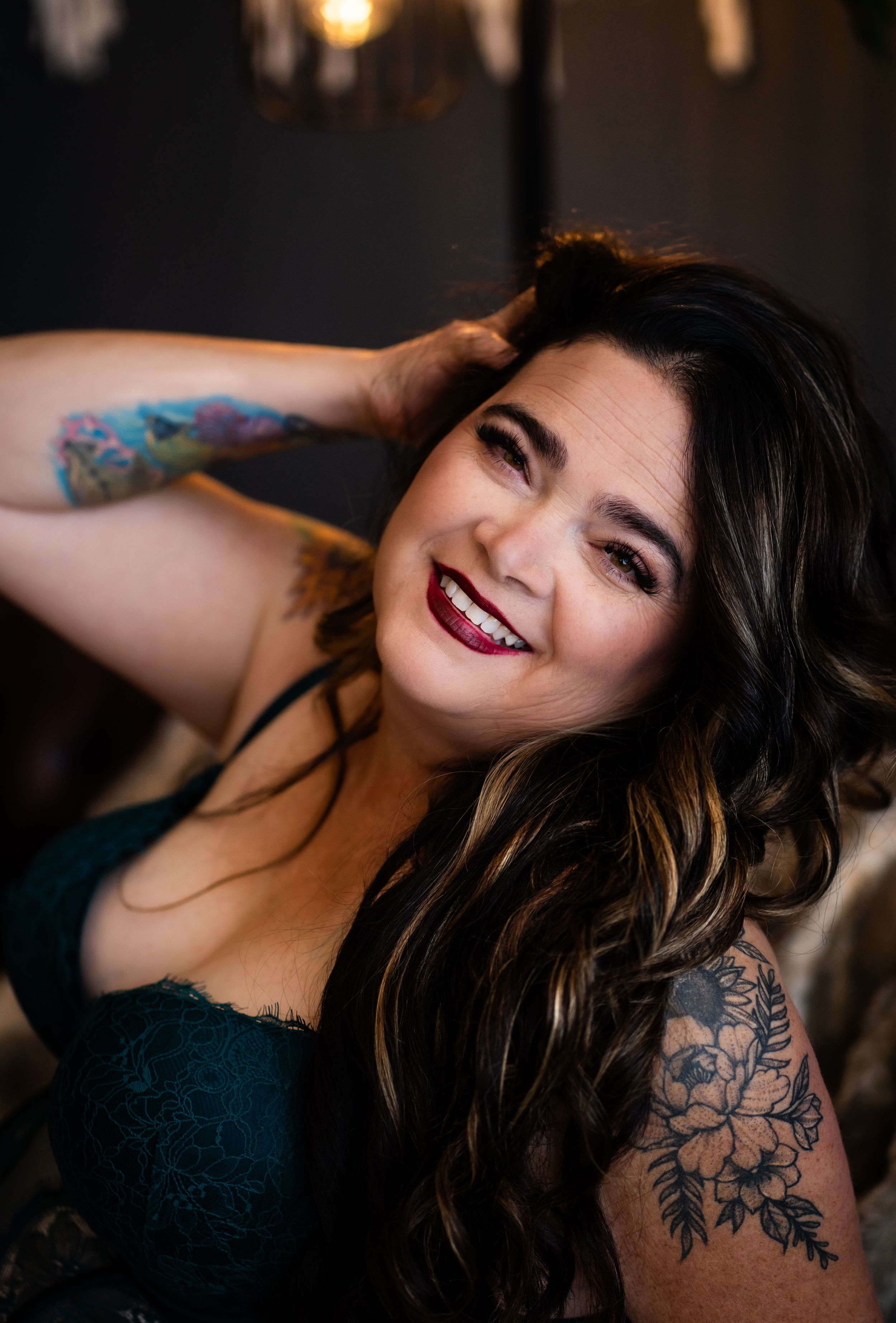 Smiling woman with dark hair, red lips, tattoos, and teal lace top poses confidently in a boudoir portrait.