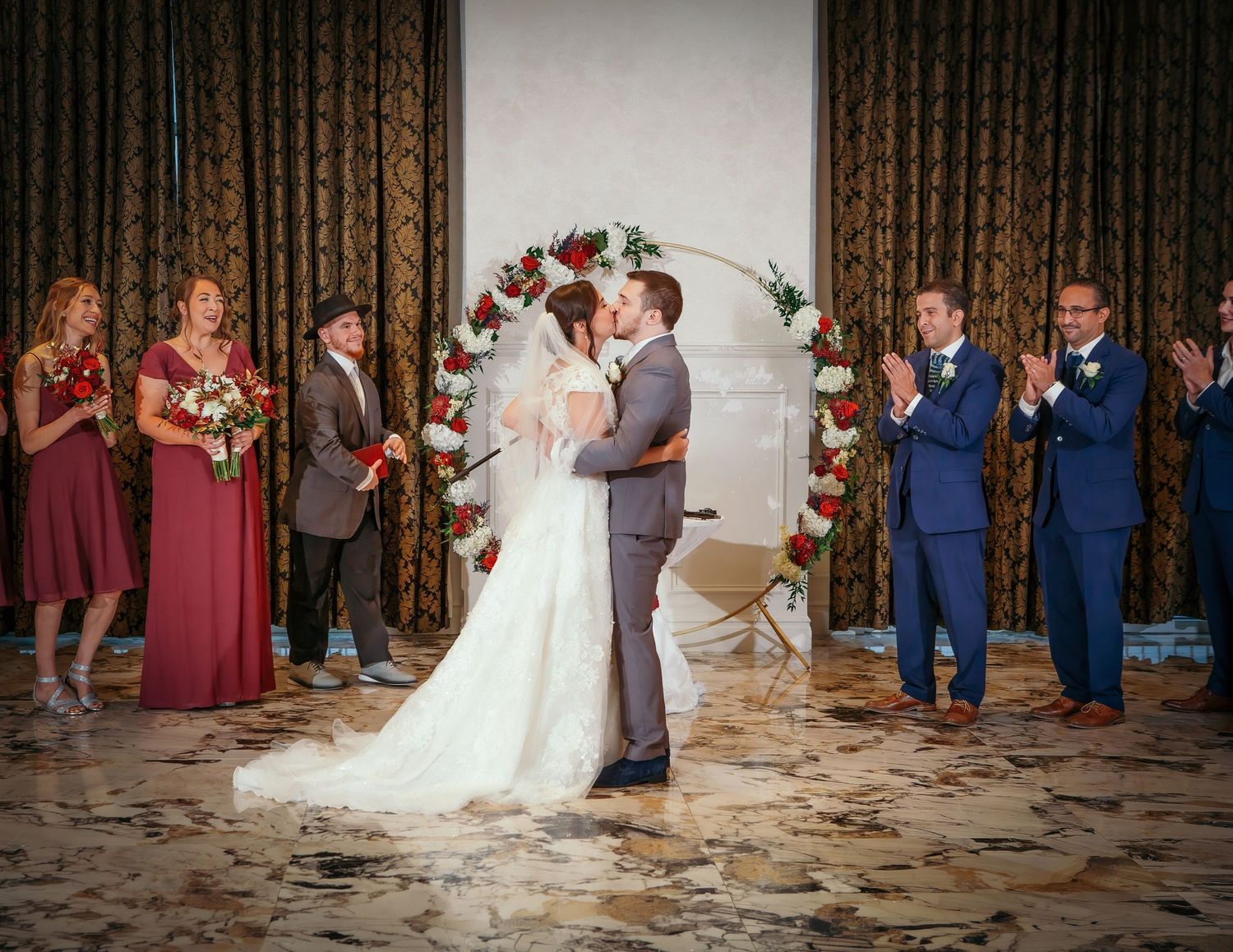 Bride and groom share first kiss at wedding ceremony with floral arch, bridesmaids in burgundy, and groomsmen applauding.