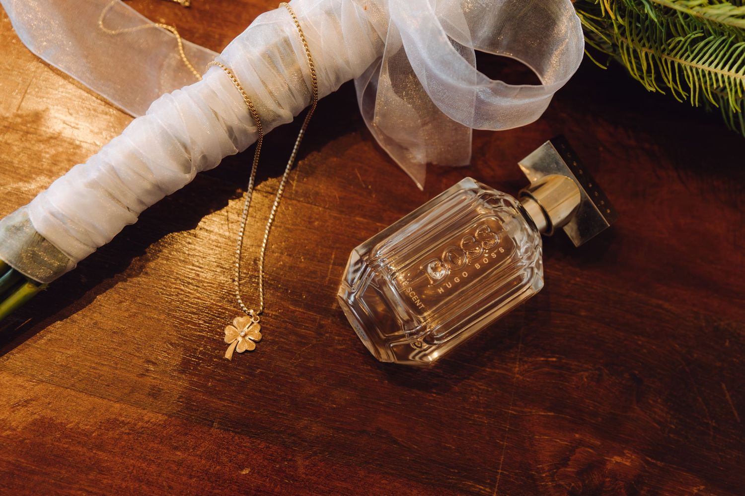 Gold necklace and perfume bottle arranged on wooden table with white tulle and green foliage.