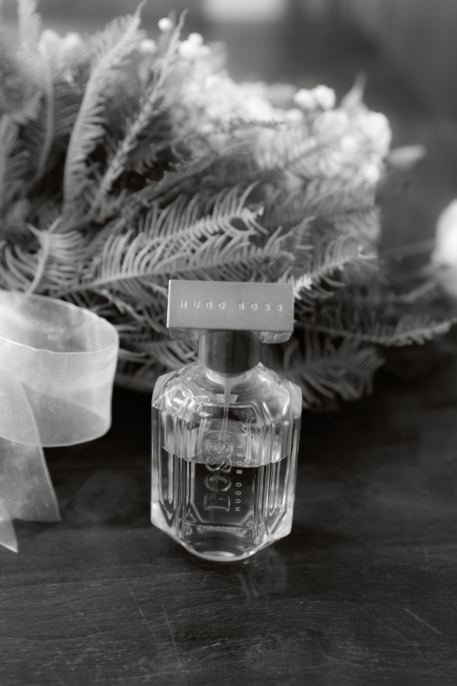 Black and white photo of Hugo Boss perfume bottle beside wedding bouquet with ferns and white ribbon.