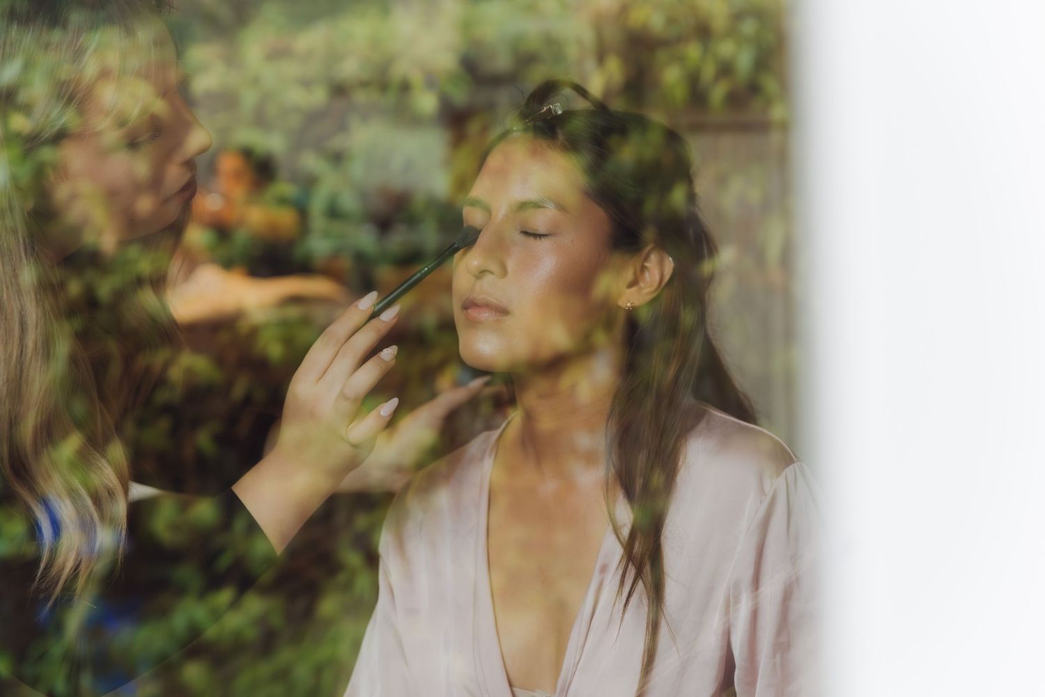 Bride getting makeup applied by artist, photographed through glass window with garden reflection.