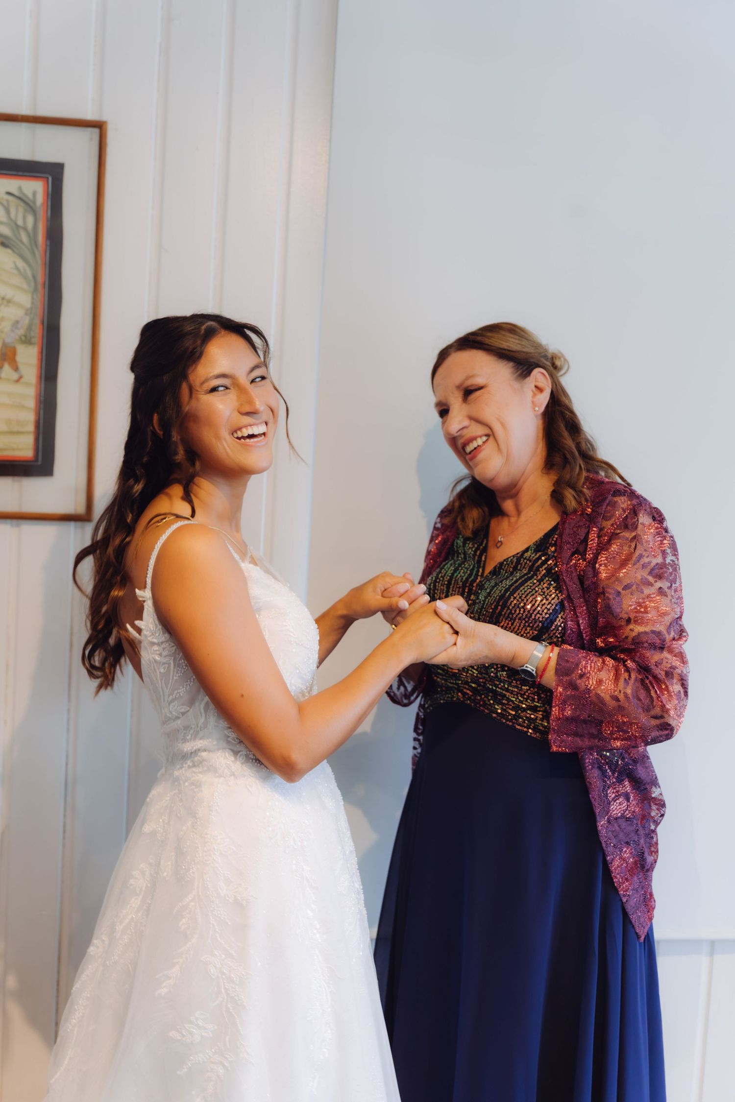 Smiling bride in white gown holding hands with older woman in navy dress and purple jacket.