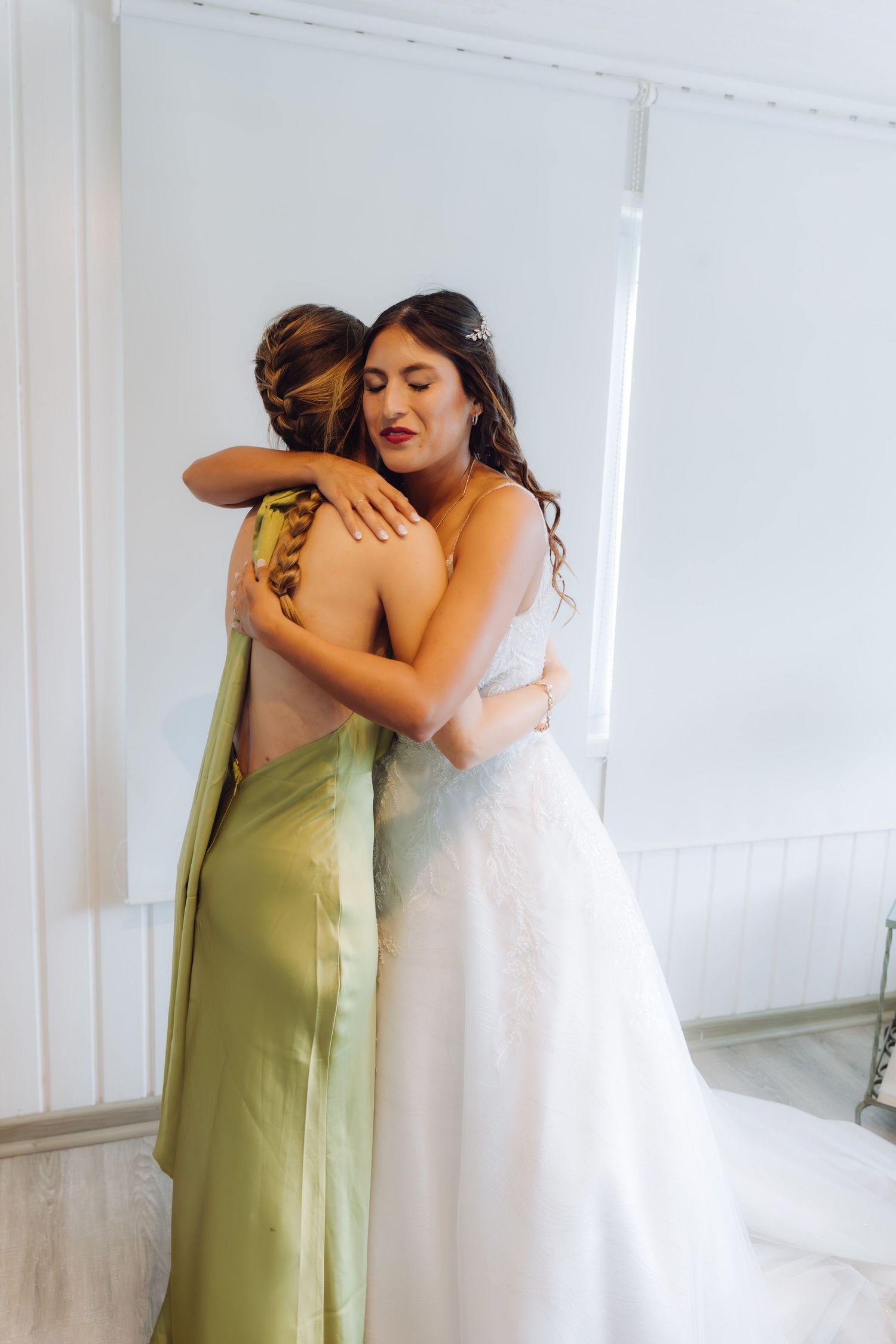 Bride in white lace gown sharing emotional hug with bridesmaid in olive green satin dress.