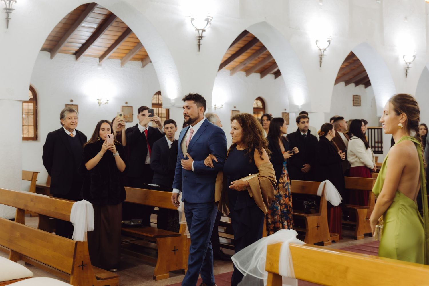 Groom in blue suit walking down church aisle with guests applauding during wedding ceremony.