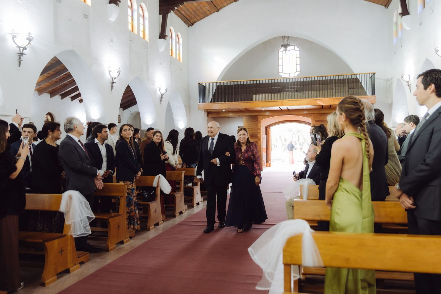 Wedding processional in white church with guests standing as bride walks down red carpet aisle.