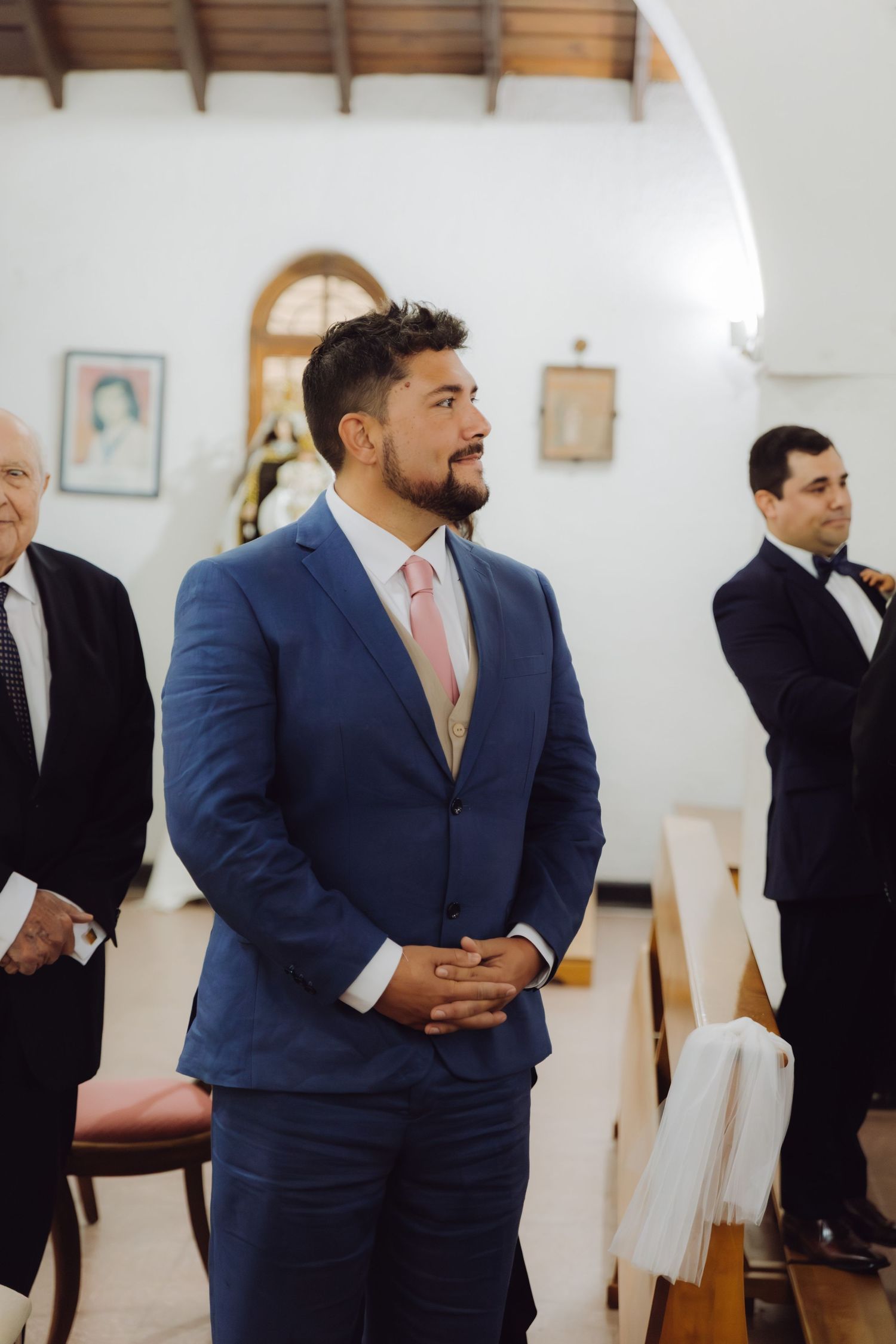 Groom in navy blue suit with pink tie smiling at altar awaiting bride in white church.