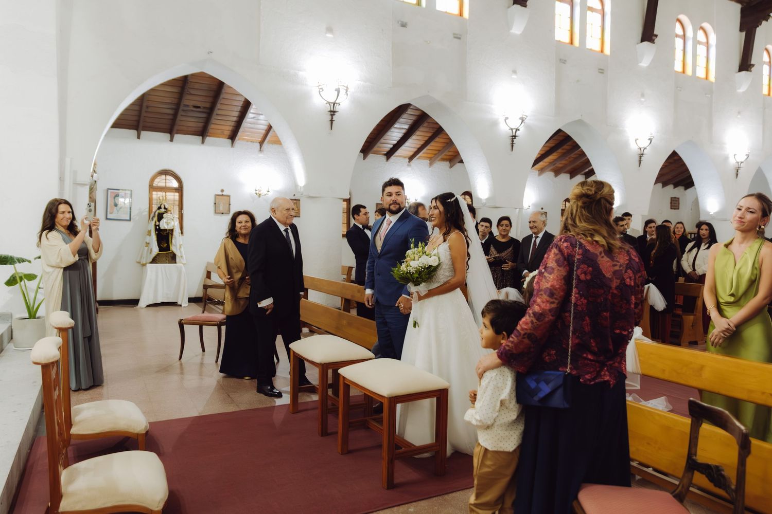 Bride walks down church aisle in white wedding gown with groom and guests watching.