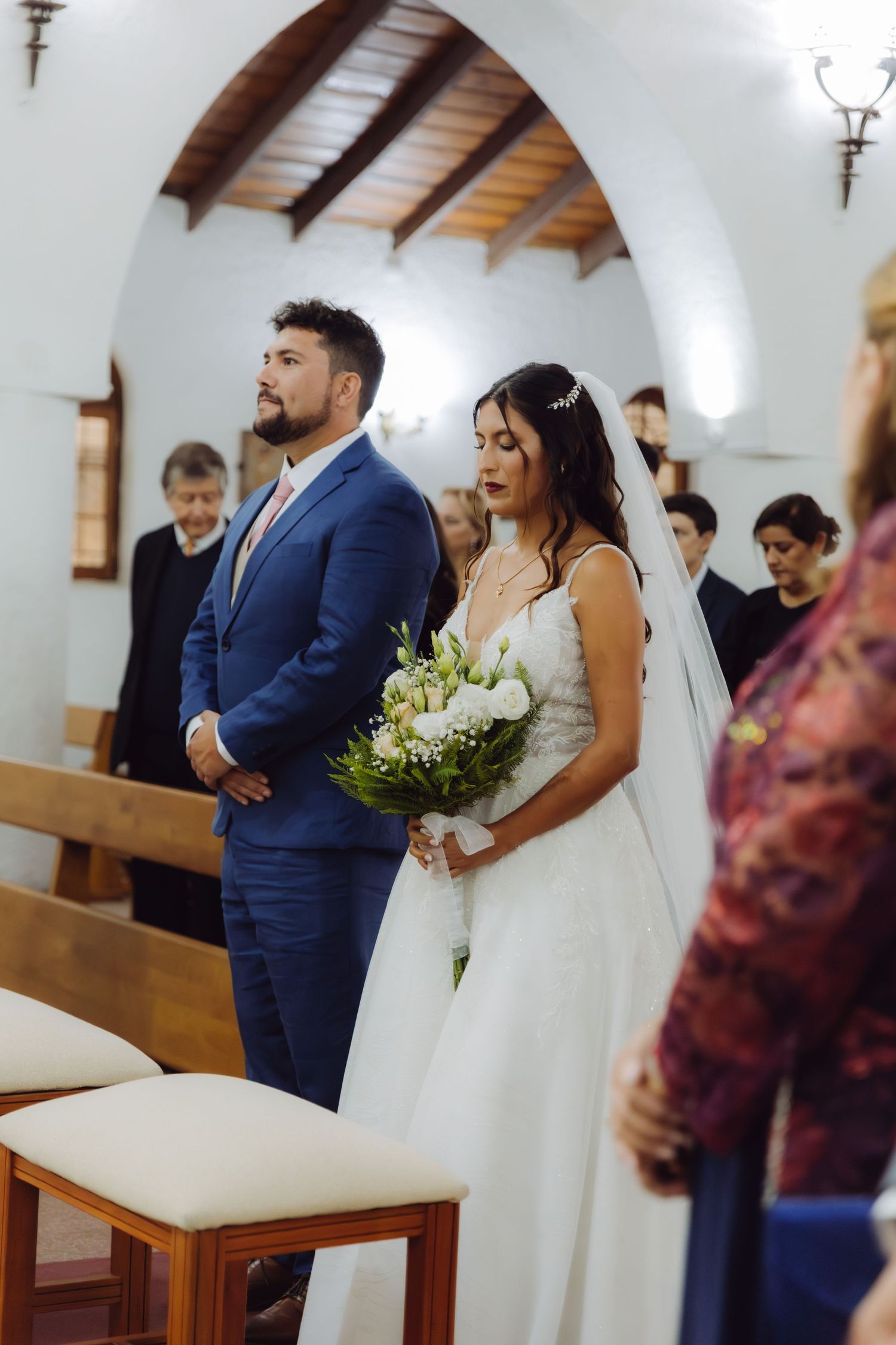 Bride holding white bouquet and groom in blue suit stand at church altar during wedding.