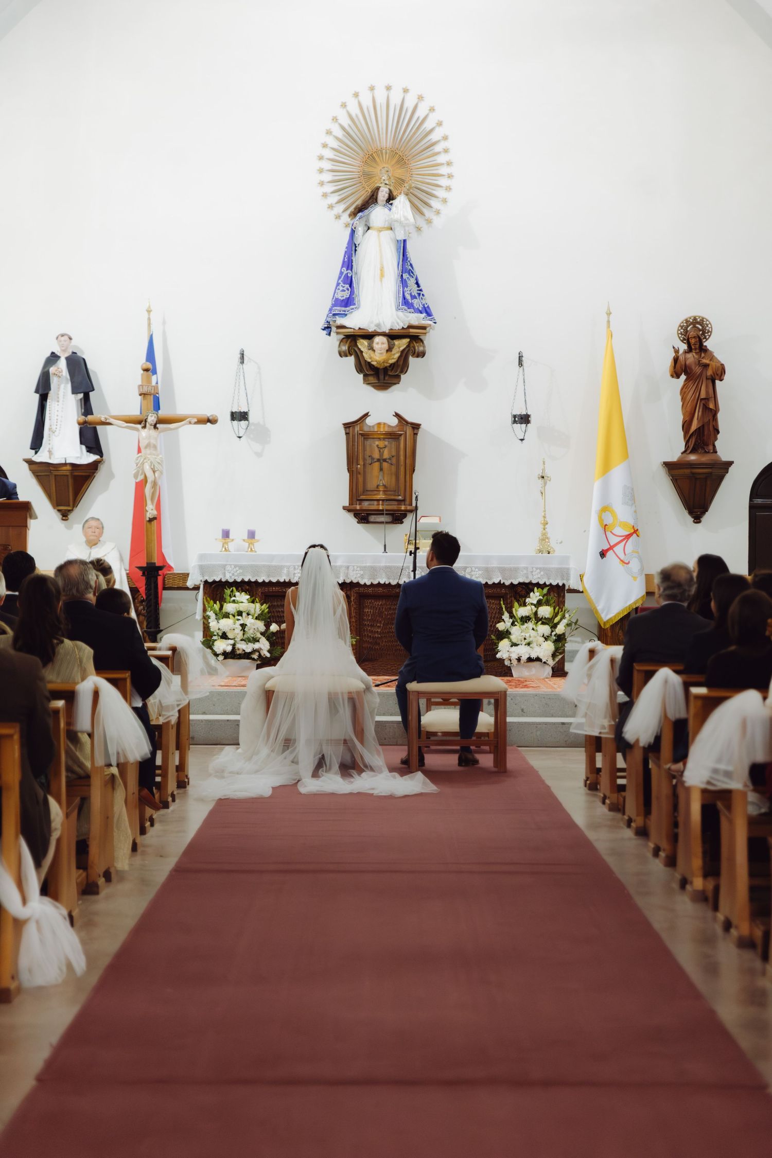 Bride and groom kneel at church altar before Virgin Mary statue during wedding ceremony.