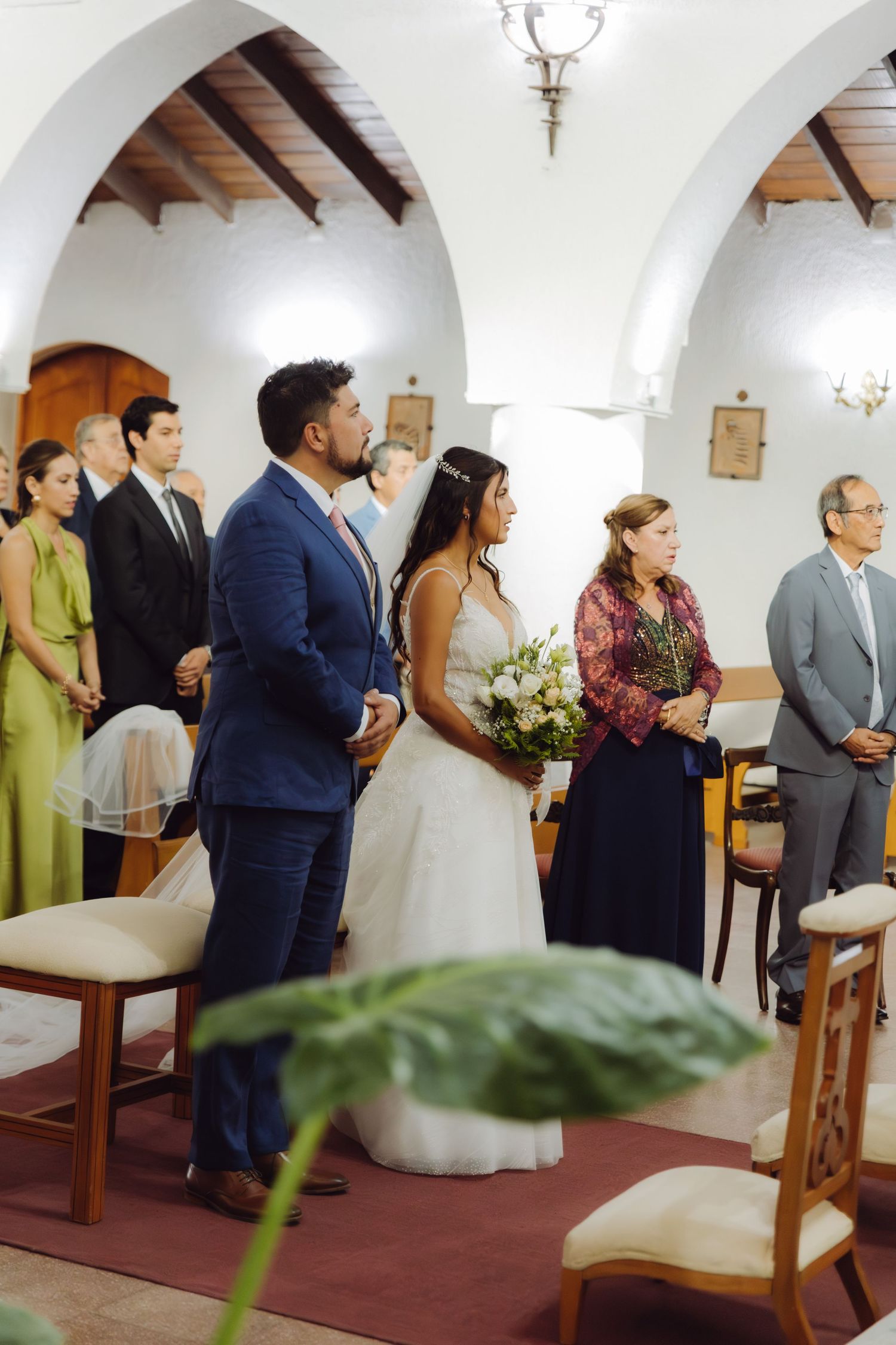 Bride with bouquet and groom in blue suit stand with family during church wedding ceremony.