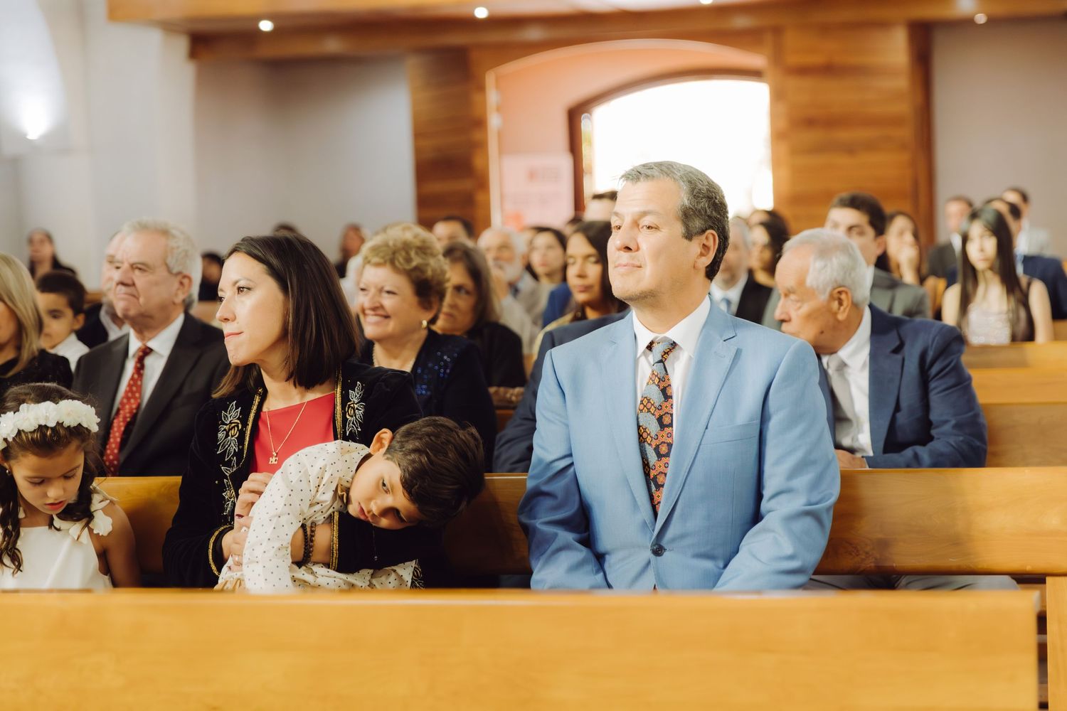 Wedding guests seated in church pews watching ceremony with man in light blue suit.