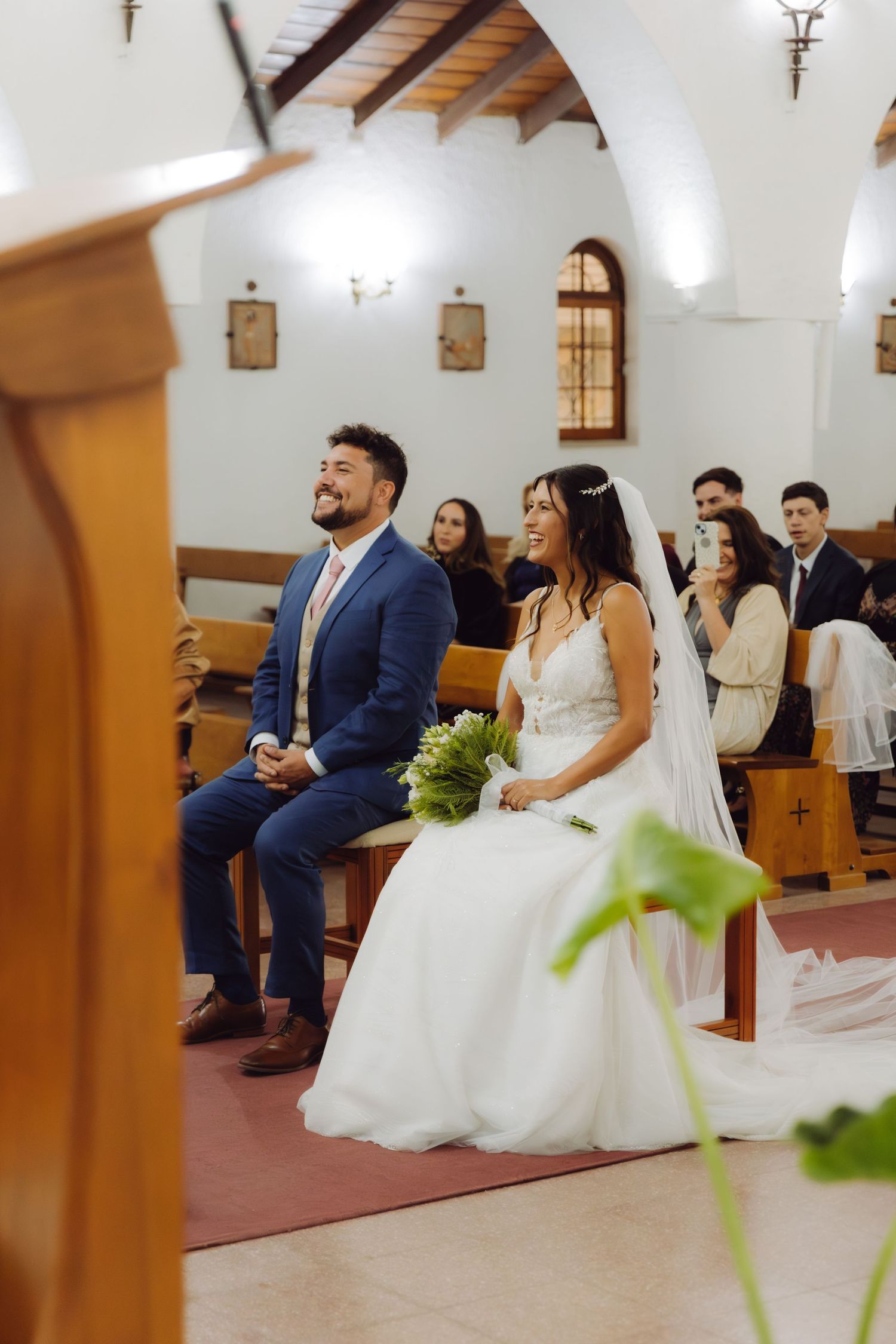 Smiling bride and groom sit together on church bench during wedding ceremony.
