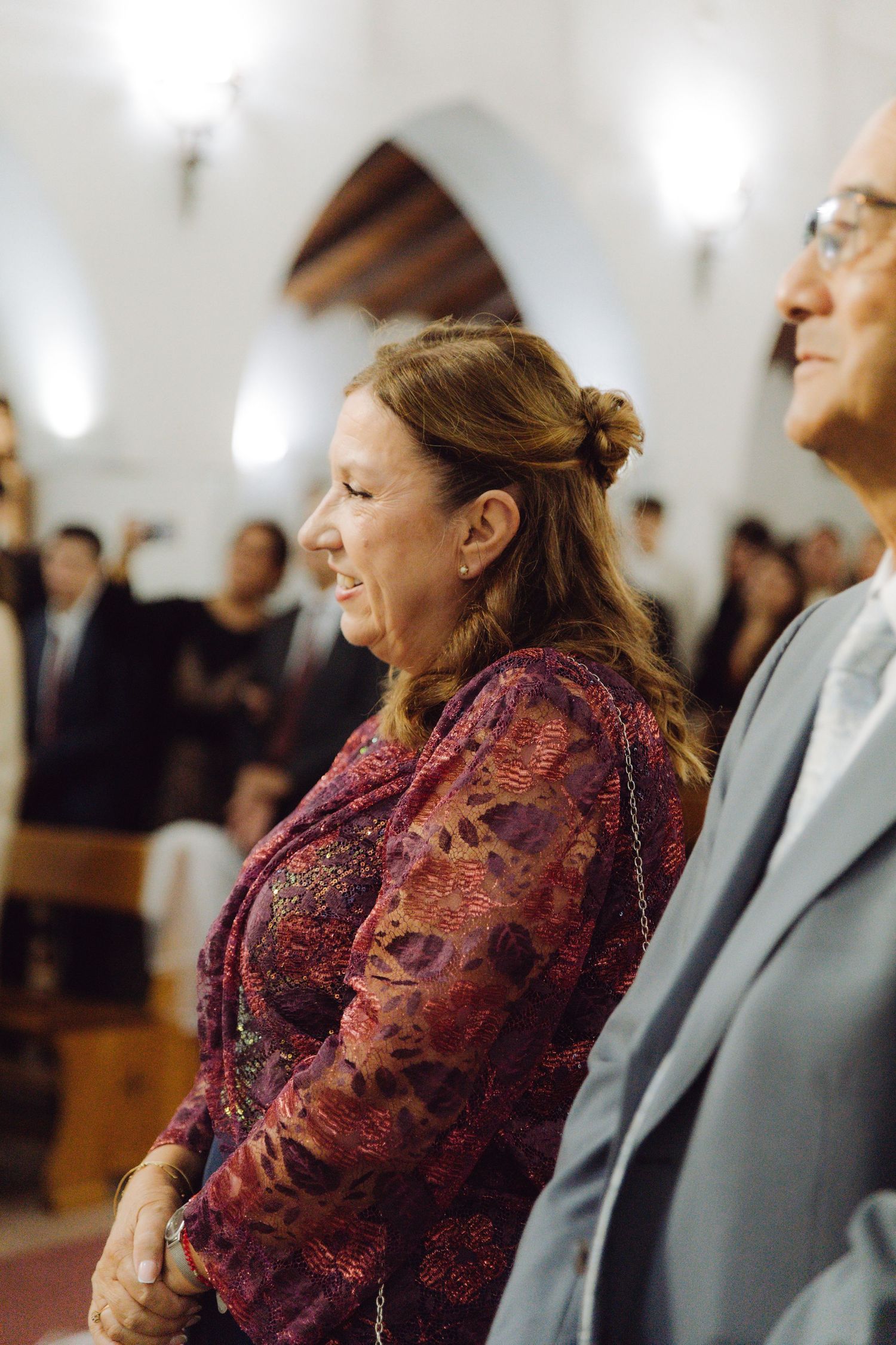 Woman in burgundy floral dress smiling warmly during a church wedding ceremony.