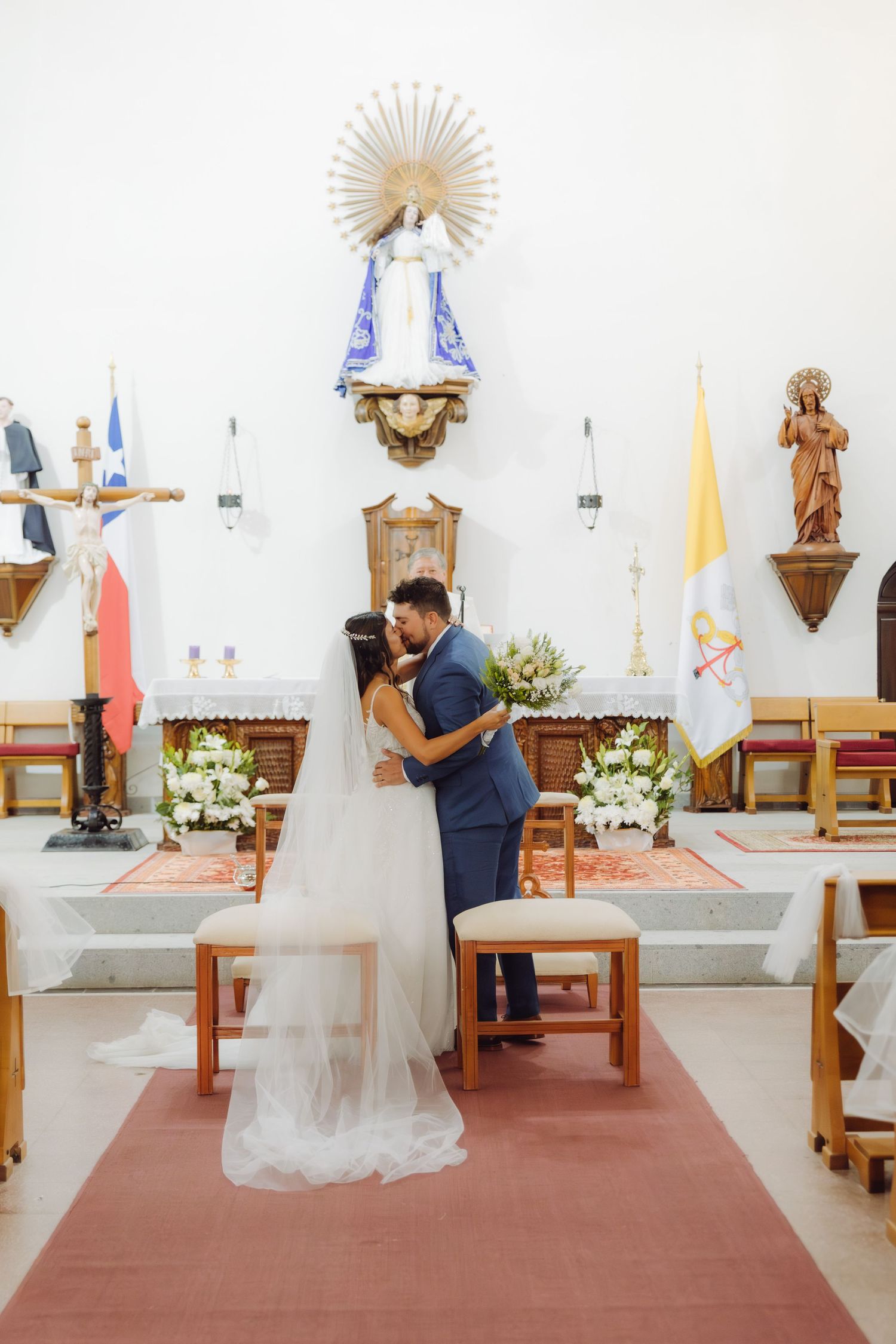 Bride and groom share first kiss at Catholic church altar during wedding ceremony with floral decorations.