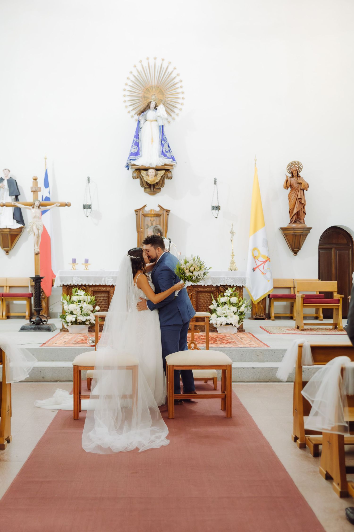 Newlyweds embrace and kiss at church altar with Vatican and Chilean flags during wedding ceremony.