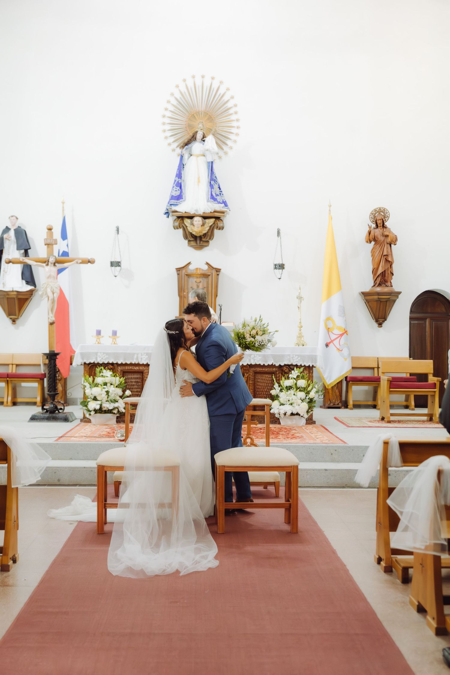Bride in white gown with veil and groom in blue suit kiss at decorated church altar.