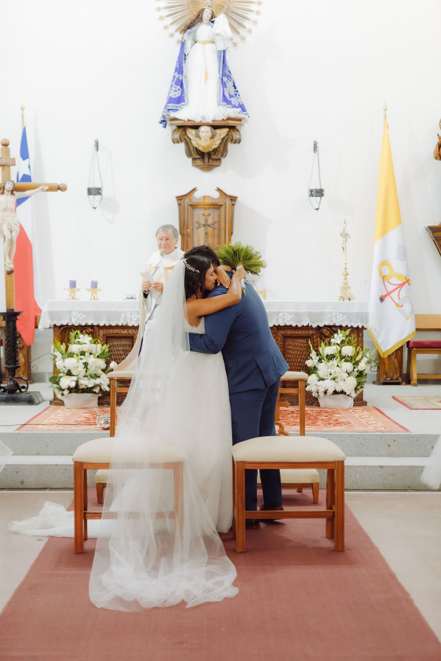 Priest watches as bride and groom share romantic kiss at Catholic church altar during ceremony.