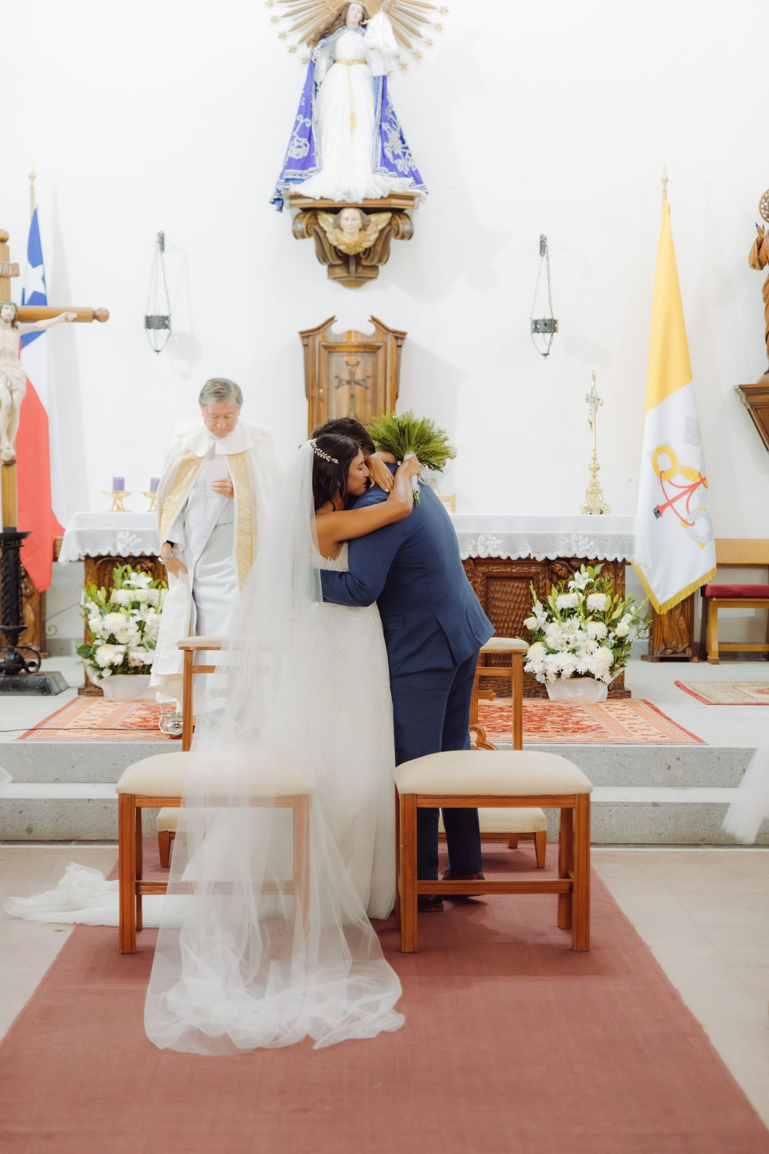 Groom embraces bride in flowing white veil at church altar while priest stands behind them.